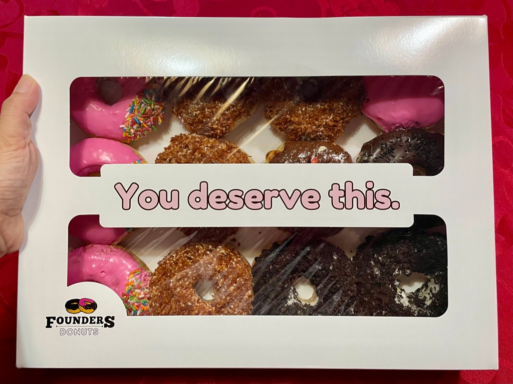 Box of 12 assorted Founders Donuts in white packaging with the message âYou deserve this.â visible on the box cover. Includes pink frosted, toasted coconut, and chocolate-flavored donuts.