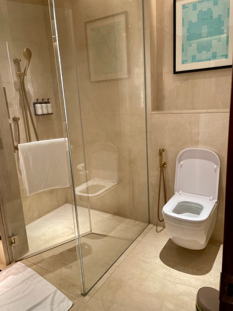 Modern bathroom at Solaire Resort North featuring a glass-enclosed shower, wall-mounted toilet, and minimalist beige marble design.
