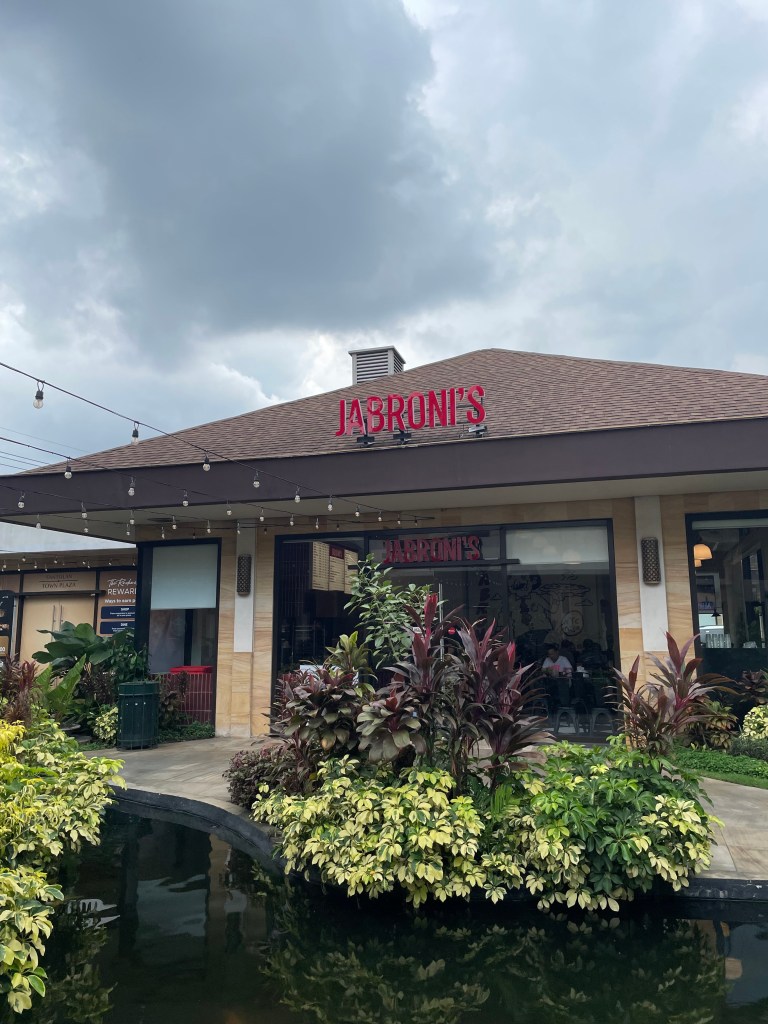 Front view of Jabroni’s New York Pizza branch at Santolan Town Plaza in Quezon City, with outdoor greenery and the restaurant sign visible under a cloudy sky.